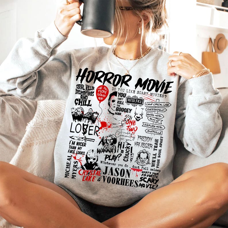 May include: A light gray sweatshirt with a graphic design featuring the words "HORROR MOVIE" and various horror movie references, including characters and phrases. The design is in black and red.