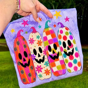 May include: A light purple t-shirt featuring a graphic of four tall, colorful pumpkins with black jack-o'-lantern faces. The pumpkins have distinct patterns including stripes with flowers, floral prints, a checkered design, and polka dots, surrounded by small stars.