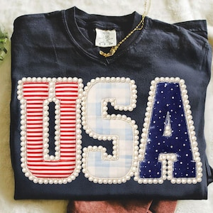 May include: Navy blue t-shirt with the word "USA" appliqued on the front. The letters are decorated with red and white stripes, blue gingham, and blue with white polka dots. Each letter is outlined with white pearls.