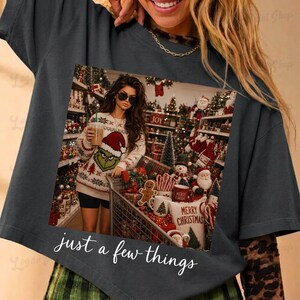 May include: A dark gray t-shirt featuring a graphic print of a person wearing a Grinch-themed sweater and sunglasses, holding a drink in a store aisle filled with Christmas decorations, with the white cursive text just a few things below.