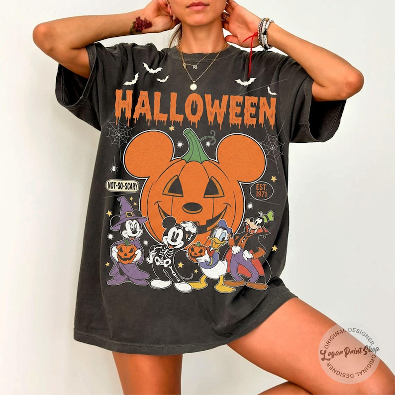 May include: Dark gray Halloween-themed t-shirt featuring a large orange pumpkin with Mickey Mouse ears and the word "HALLOWEEN" above. Below, Mickey, Minnie, Donald, and Goofy are dressed in Halloween costumes. The shirt has the text "NOT-SO-SCARY" and "EST. 1971".