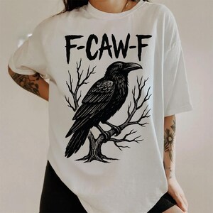 May include: White t-shirt with a black graphic of a raven perched on a tree branch. The text "F-CAW-F" is above the raven. The shirt has short sleeves and a relaxed fit.