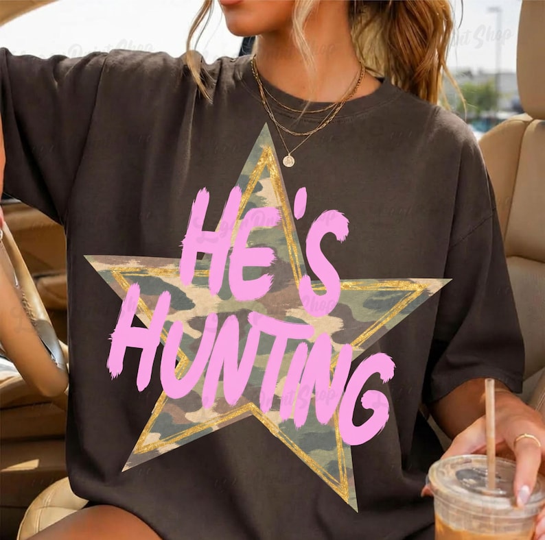 May include: A dark brown short-sleeved t-shirt featuring a large camouflage-patterned star graphic. The text "HE'S HUNTING" is printed across the star in a bold, bright pink brushstroke font. The star has a thin, distressed gold-colored outline.