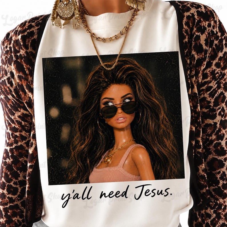 May include: A white t-shirt featuring a graphic print of a doll with long brown hair wearing sunglasses and a pink top. The text "y'all need Jesus." is printed below the graphic. The shirt is worn under a brown and black leopard print cardigan.