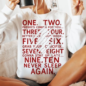 May include: White sweatshirt with the text "ONE, TWO, FREDDY'S COMING FOR YOU..." in red. The design includes claw marks. The sweatshirt is a casual garment.