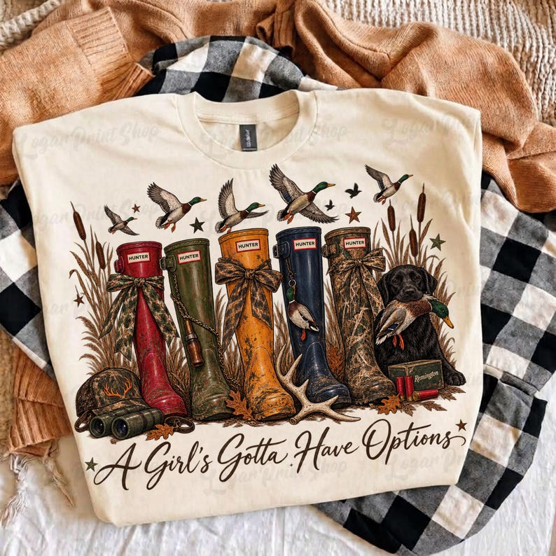 May include: A cream-colored t-shirt featuring a graphic of five rubber boots in red, olive, yellow, navy, and camouflage, alongside a black dog holding a duck, cattails, and the text A Girl's Gotta Have Options in a cursive black font.