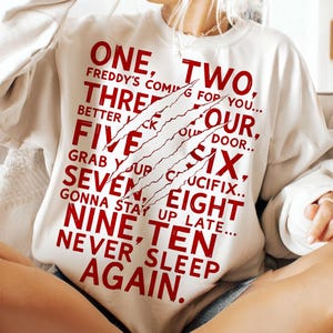 May include: Cream-colored sweatshirt with the text "ONE, TWO, THREE, FOUR, FIVE, SIX, SEVEN, EIGHT, NINE, TEN, NEVER SLEEP AGAIN" in red, with claw marks. The sweatshirt is a casual style.