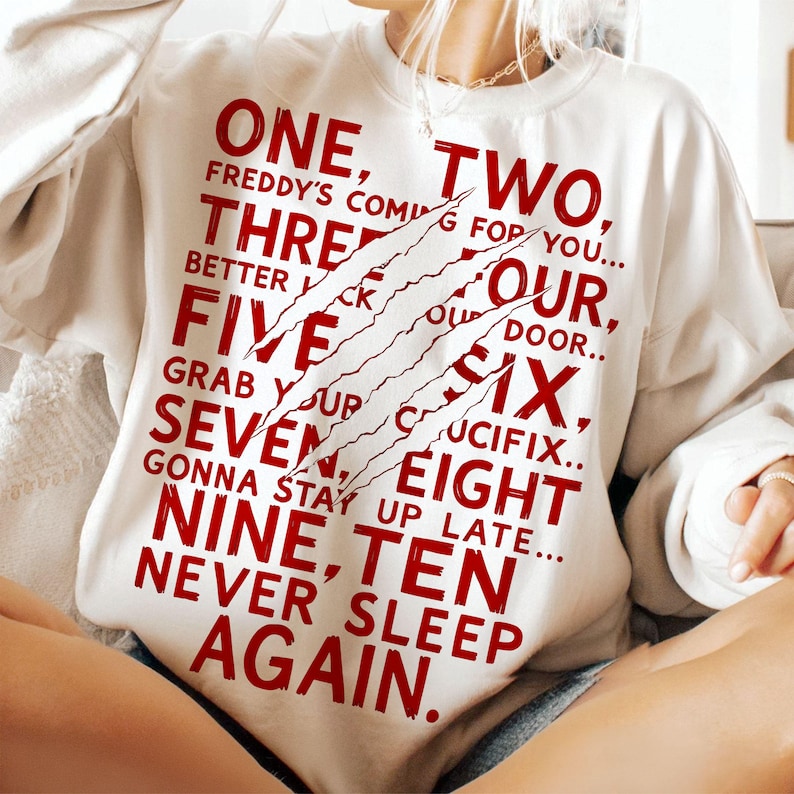 May include: Cream-colored sweatshirt with the text "ONE, TWO, THREE, FOUR, FIVE, SIX, SEVEN, EIGHT, NINE, TEN, NEVER SLEEP AGAIN" in red, with claw marks. The sweatshirt is a casual style.