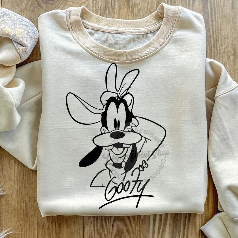 May include: A cream-colored sweatshirt with a black and white illustration of Goofy, a cartoon dog character, wearing a hat. The text "Goofy" is written in black below the illustration.