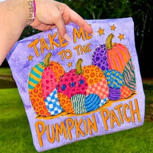Pode incluir: Uma t-shirt roxa clara com três abóboras com secções coloridas e estampadas, incluindo padrão de leopardo, riscas e estrelas. O texto "TAKE ME TO THE PUMPKIN PATCH" está impresso em letras cor de laranja acima e abaixo das abóboras.