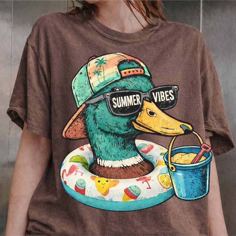 May include: Brown t-shirt featuring a cartoon duck wearing sunglasses that read "Summer Vibes." The duck is wearing a hat and a swim ring. It is holding a blue bucket with sand and a red shovel. The shirt has a vintage, distressed look.
