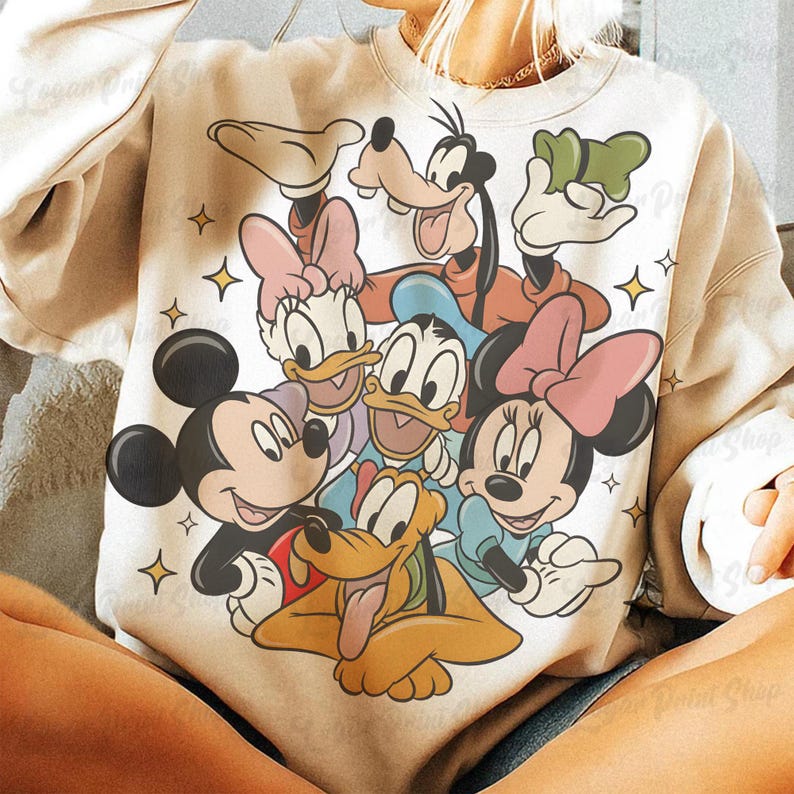 May include: Cream-colored sweatshirt featuring a colorful graphic of Disney characters Mickey Mouse, Minnie Mouse, Donald Duck, Daisy Duck, Goofy, and Pluto. The design includes star accents. The sweatshirt is a casual style.