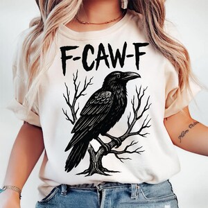 May include: Cream-colored t-shirt featuring a black illustration of a raven perched on a tree branch. The text "F-CAW-F" is printed above the raven in bold, black letters. The design is centered on the shirt.