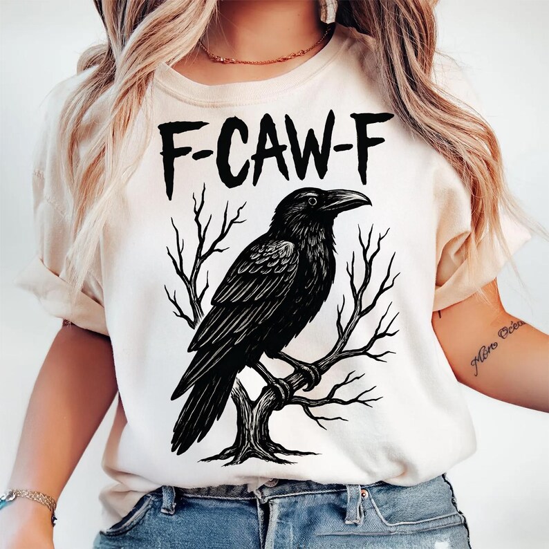 May include: Cream-colored t-shirt featuring a black illustration of a raven perched on a tree branch. The text "F-CAW-F" is printed above the raven in bold, black letters. The design is centered on the shirt.