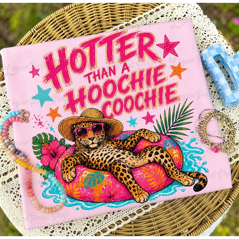 Puede incluir: Camiseta rosa con la frase "Hotter Than A Hoochie Coochie" en letras rosas brillantes. Un leopardo con gafas de sol y sombrero de paja descansa sobre un flotador inflable rosa. Estrellas y flores tropicales decoran la camiseta.