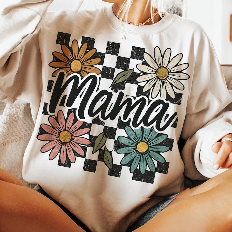 May include: White sweatshirt with a checkered pattern and colorful daisies. The word "Mama" is written in a retro font across the center of the design.
