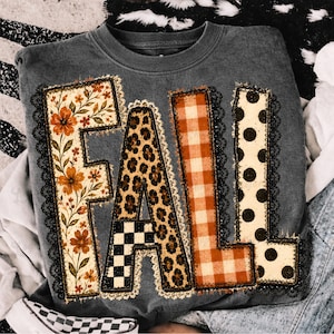 May include: A charcoal gray crewneck sweatshirt featuring the word FALL in large block letters. Each letter has a unique pattern: floral, leopard print, orange gingham, and black polka dots, all outlined with black lace trim detailing.