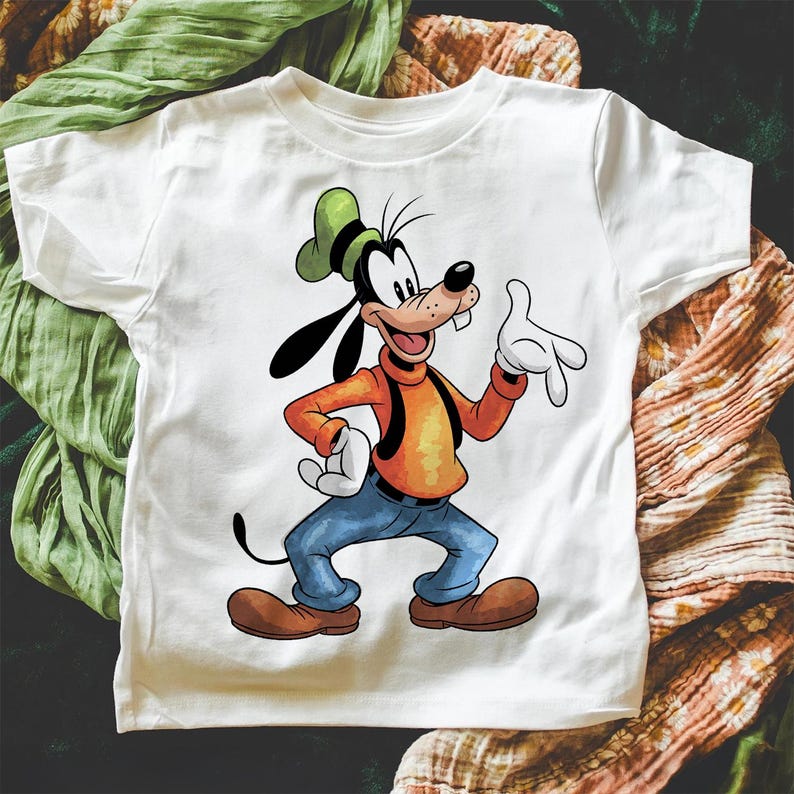 May include: White t-shirt with a cartoon image of Goofy, a dog character from Disney, wearing a green hat, orange shirt, and blue jeans. Goofy is smiling and waving.