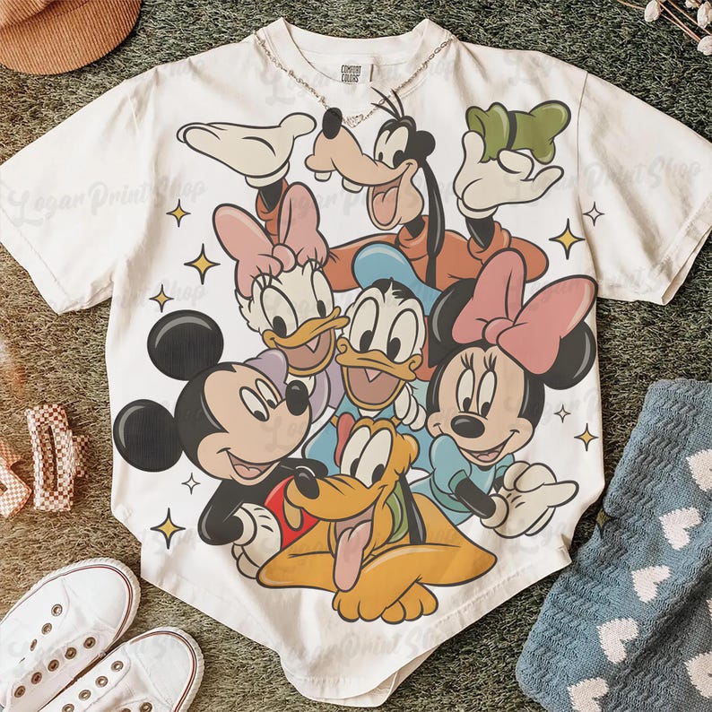 May include: White t-shirt featuring a colorful graphic of Disney characters including Mickey Mouse, Minnie Mouse, Donald Duck, Daisy Duck, Goofy, and Pluto. The design is surrounded by yellow stars. The shirt is on a green surface with shoes and a hat.