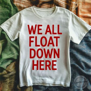 May include: White t-shirt with the red text "WE ALL FLOAT DOWN HERE". The shirt has short sleeves and a crew neck. The design is centered on the front of the shirt. The shirt is made of a soft material.