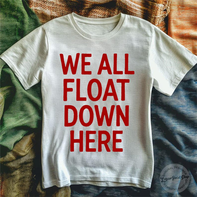 May include: White t-shirt with the red text "WE ALL FLOAT DOWN HERE". The shirt has short sleeves and a crew neck. The design is centered on the front of the shirt. The shirt is made of a soft material.