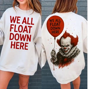 May include: White sweatshirt with the text "WE ALL FLOAT DOWN HERE" in red. The back features a graphic of a clown holding a red balloon with the same text. The clown has red hair and a sinister smile.