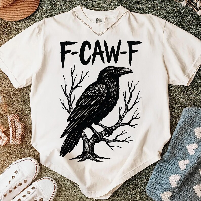 May include: Cream-colored t-shirt with a black raven illustration perched on a tree branch. The text "F-CAW-F" is above the raven. The shirt is a Comfort Colors brand.