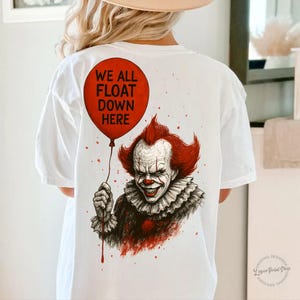 May include: White t-shirt featuring a graphic of a menacing clown holding a red balloon with the text "WE ALL FLOAT DOWN HERE". The clown has red hair and a ruffled collar.