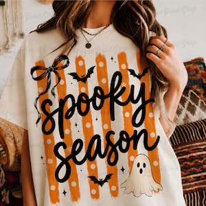 Op de afbeelding: Een cr&egrave;mekleurig t-shirt met een grafische print met de tekst "spooky season" in zwart handschrift. Het ontwerp bevat verticale oranje strepen met witte stippen, een zwarte strik, drie zwarte vleermuizen en een kleine witte spookillustratie.