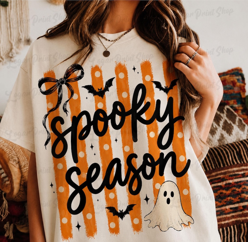 Op de afbeelding: Een cr&egrave;mekleurig t-shirt met een grafische print met de tekst "spooky season" in zwart handschrift. Het ontwerp bevat verticale oranje strepen met witte stippen, een zwarte strik, drie zwarte vleermuizen en een kleine witte spookillustratie.