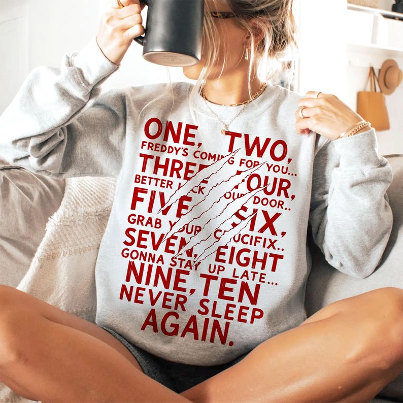 May include: A light gray sweatshirt with the text "ONE, TWO, FREDDY'S COMING FOR YOU..." in red. The design includes claw marks. The sweatshirt is a casual garment.