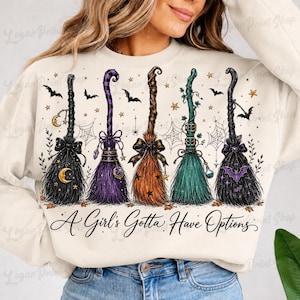 May include: A cream-colored sweatshirt featuring a graphic of five distinct witch brooms in black, purple, orange, green, and dark purple. The design includes stars, spiderwebs, bats, and the text A Girl's Gotta Have Options in black script.