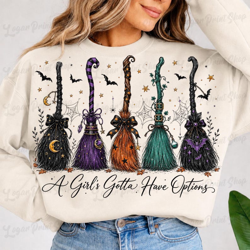 May include: A cream-colored sweatshirt featuring a graphic of five distinct witch brooms in black, purple, orange, green, and dark purple. The design includes stars, spiderwebs, bats, and the text A Girl's Gotta Have Options in black script.