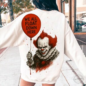 May include: Cream-colored sweatshirt featuring a graphic of a menacing clown holding a red balloon. The balloon has the text "WE ALL FLOAT DOWN HERE". The clown has red hair and a ruffled collar. The design is a horror-themed graphic.