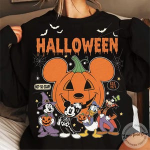 May include: Black Halloween sweatshirt featuring a large orange pumpkin with Mickey Mouse ears and a smiling face. The word "HALLOWEEN" is above the pumpkin, with bats and cartoon characters below. Text includes "NOT-SO-SCARY" and "EST. 1971."