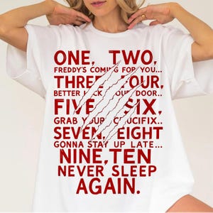 May include: White t-shirt with red text and claw marks. The text reads "ONE, TWO, FREDDY'S COMING FOR YOU... THREE, FOUR, BETTER LOCK YOUR DOOR... FIVE, SIX, GRAB YOUR CRUCIFIX... SEVEN, EIGHT, GONNA STAY UP LATE... NINE, TEN, NEVER SLEEP AGAIN."