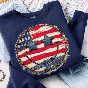 May include: Navy blue sweatshirt featuring a distressed American flag smiley face design. The smiley face has denim star eyes and a stitched denim border. The sweatshirt is folded on top of a pair of blue jeans.