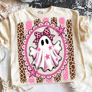 May include: A cream-colored t-shirt featuring a central graphic of a white ghost wearing a pink polka-dot bow. The ghost is inside a pink scalloped oval frame decorated with pink stars, set against a background of vertical leopard print stripes.