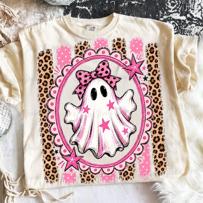 May include: A cream-colored t-shirt featuring a central graphic of a white ghost wearing a pink polka-dot bow. The ghost is inside a pink scalloped oval frame decorated with pink stars, set against a background of vertical leopard print stripes.