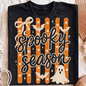 Op de afbeelding: Een zwart t-shirt met een grafische print van verticale oranje en zwarte strepen, witte stippen en de tekst "spooky season" in zwart schrift. Het ontwerp bevat een witte strik, drie witte vleermuizen en een witte spookillustratie.