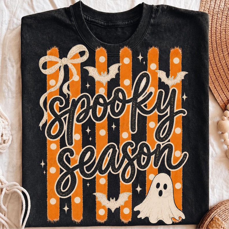 Op de afbeelding: Een zwart t-shirt met een grafische print van verticale oranje en zwarte strepen, witte stippen en de tekst "spooky season" in zwart schrift. Het ontwerp bevat een witte strik, drie witte vleermuizen en een witte spookillustratie.