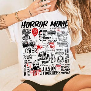 May include: White t-shirt with a graphic design featuring horror movie characters and phrases. The design includes text like "HORROR MOVIE", "SERIAL KILLER", and "FREDDY KRUEGER". The shirt has a crew neck and short sleeves.