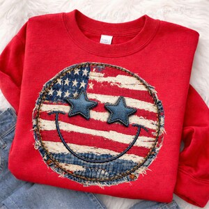 May include: Red sweatshirt featuring a distressed American flag smiley face design. The smiley face has denim star eyes and a stitched denim border. The design is centered on the front of the sweatshirt.