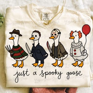 Może przedstawiać: Kremowy t-shirt z grafiką przedstawiającą cztery gęsi przebrane za postacie z horrorów. Poniżej gęsi znajduje się napis "just a spooky goose" czcionką imitującą pismo odręczne. Koszulka ma okrągły dekolt i metkę marki Comfort Colors.