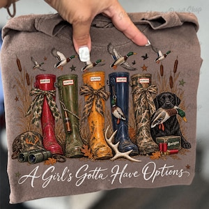 May include: A brown t-shirt featuring a graphic of four Hunter rain boots in red, olive, yellow, and camouflage, a black Labrador retriever holding a duck, binoculars, a cap, and the text A Girl's Gotta Have Options in white script.