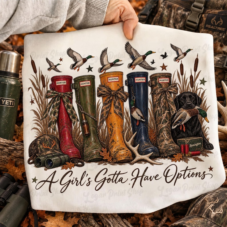 May include: A white t-shirt featuring a graphic print of five tall rubber boots in red, green, yellow, blue, and camouflage, alongside a black dog holding a duck, binoculars, and cattails. The text reads A Girl's Gotta Have Options.