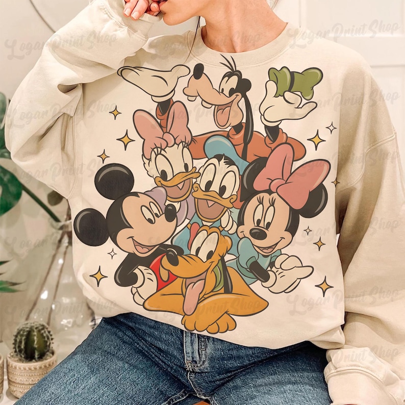 May include: A beige sweatshirt featuring a colorful graphic of Disney characters including Mickey Mouse, Minnie Mouse, Donald Duck, Daisy Duck, Goofy, and Pluto. The design is surrounded by yellow star accents.