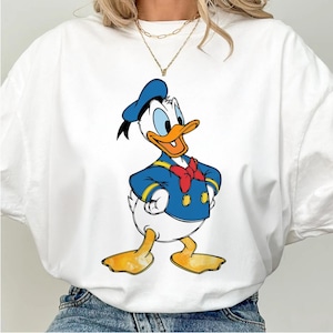 May include: White t-shirt with a cartoon image of Donald Duck wearing a blue sailor outfit and a red bow tie.