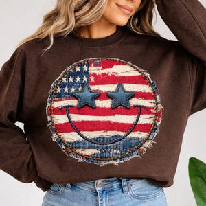May include: A brown sweatshirt featuring a distressed American flag smiley face design. The smiley face has blue star eyes and a stitched denim border. The sweatshirt is a casual style.