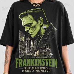 May include: A black t-shirt featuring a large graphic print of Frankenstein's monster in profile above a laboratory scene with a scientist. The text reads FRANKENSTEIN THE MAN WHO MADE A MONSTER in green lettering at the bottom of the graphic design.